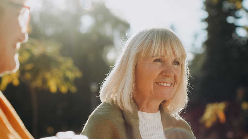 Senior woman smiling, enjoying conversation with a friend outdoors on a sunny day.